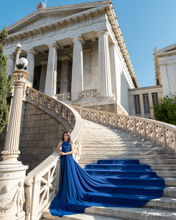 Young girl in a royal blue satin flying dress smiling on the ornate marble staircase of the National Library of Athens, with the long trailing fabric cascading elegantly down the steps.